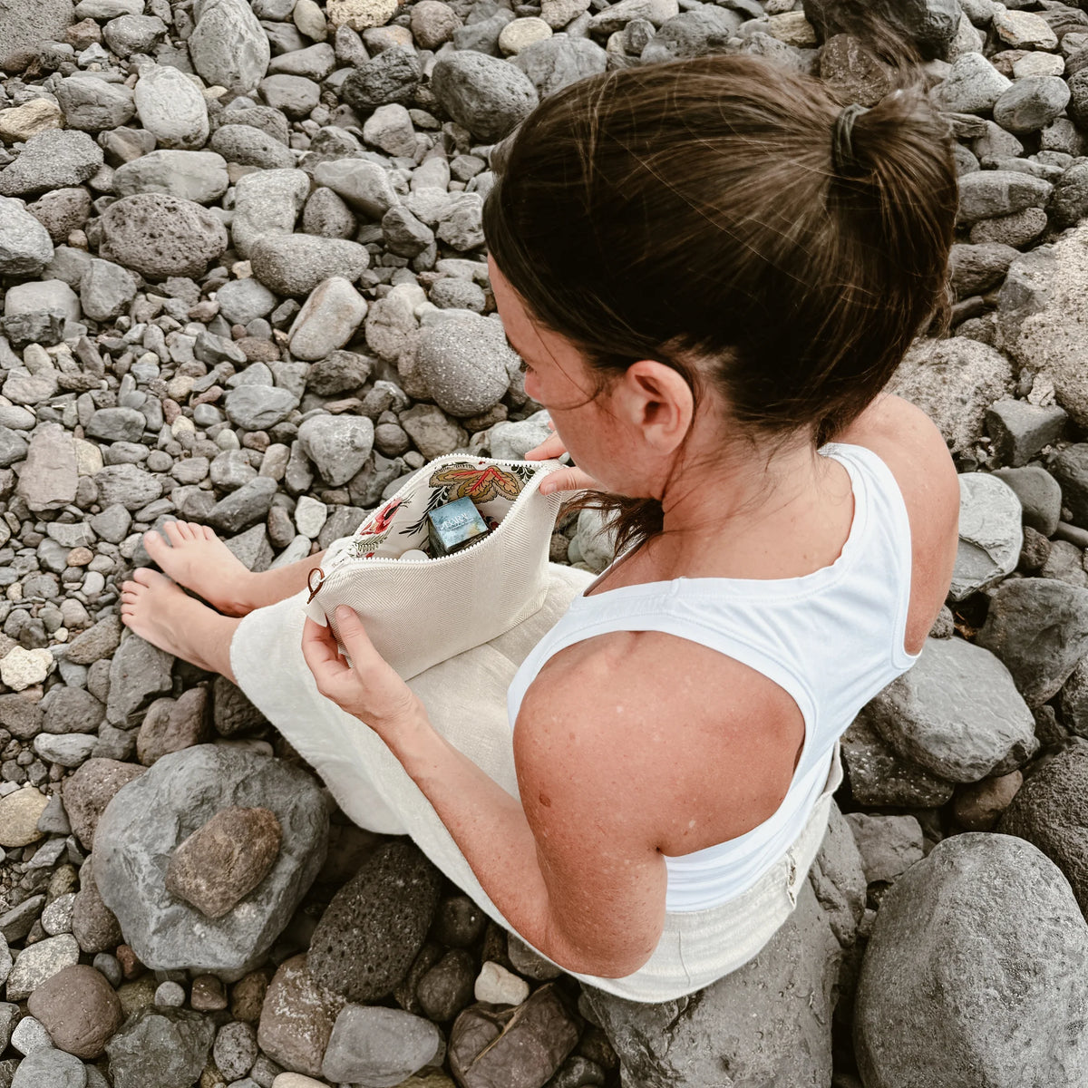 Woman sitting on rocks opening the BARAU Carry the Calm cosmetic bag